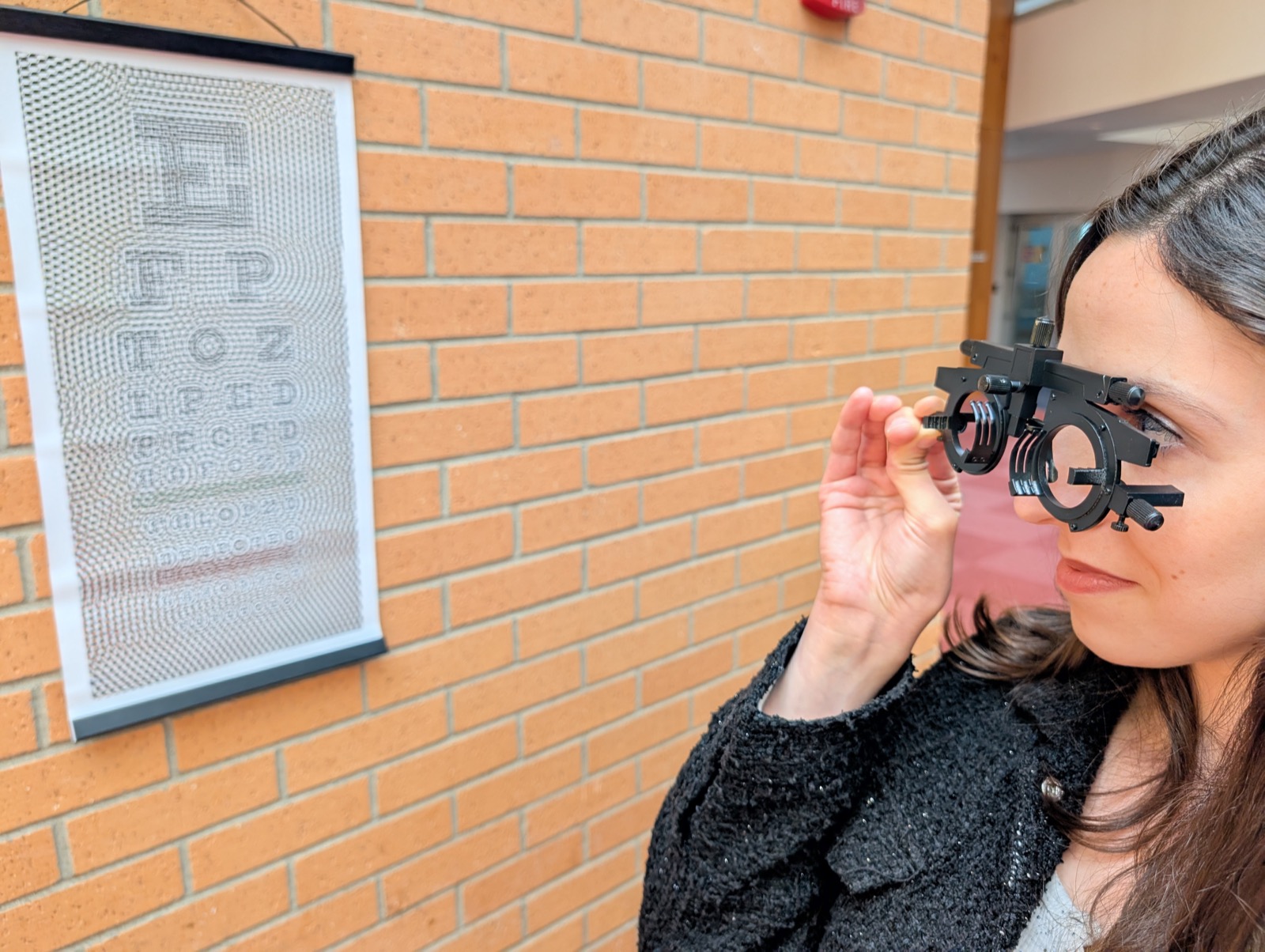 A person wearing a trial lens frame, pulling focus on a Snellen-style eye chart hung on a brick wall.